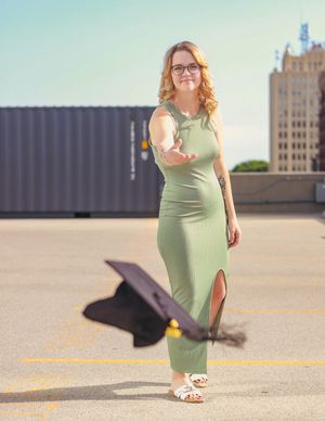 Woman in mint green fitted dress and glasses stands on urban rooftop with graduation cap flying through the air.