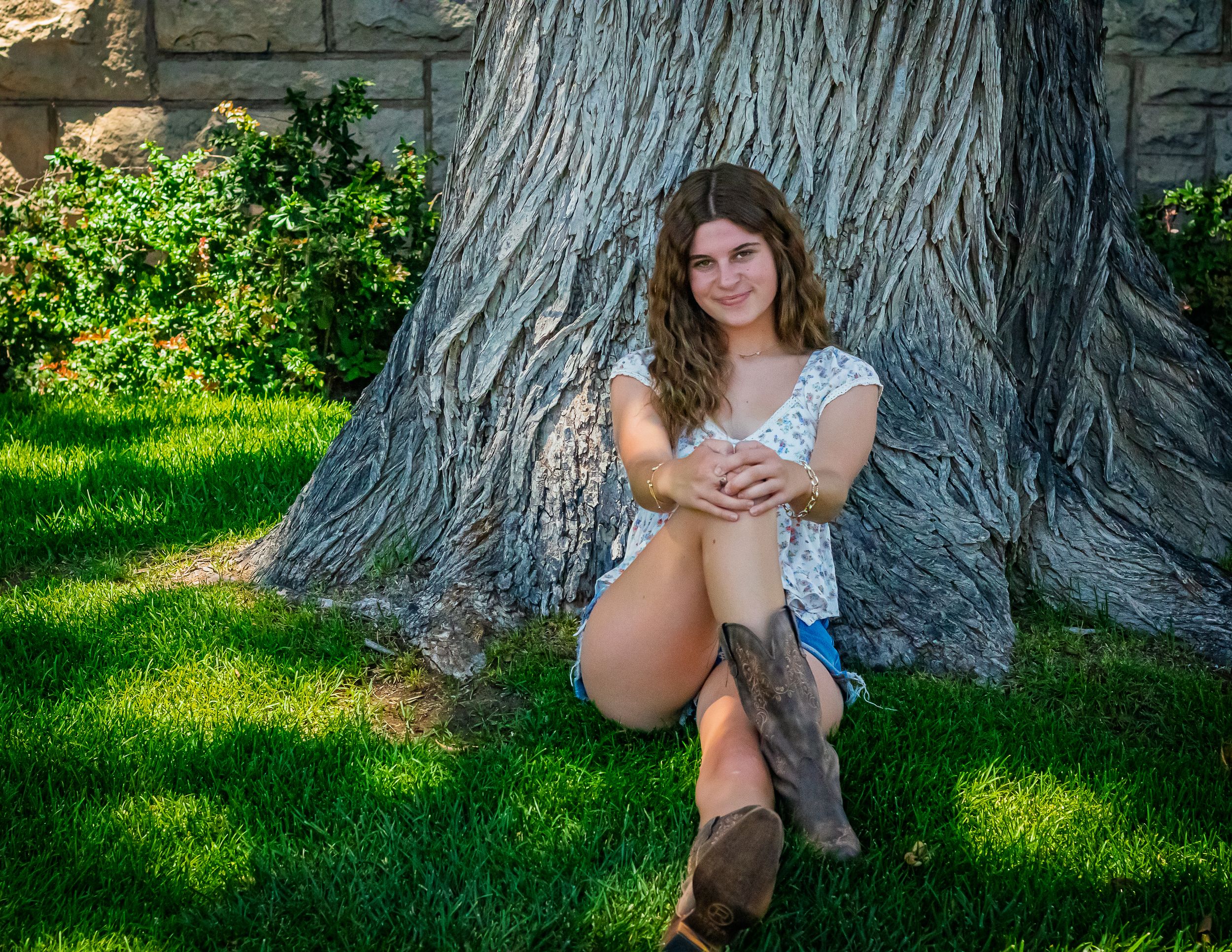 A summery outdoor portrait by a large tree trunk, with green grass and soft lighting creating a natural setting.