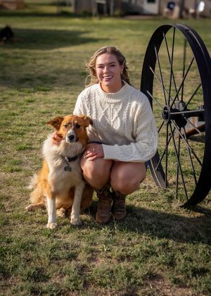 A person wearing a white sweater kneels on grass with a collie-type dog next to an antique wagon wheel.