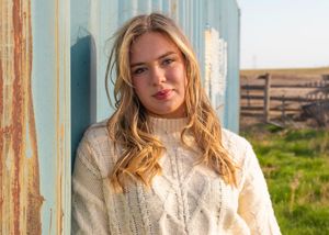 Person wearing a cozy cream cable knit sweater stands against rustic blue wall in warm sunlight with countryside view.