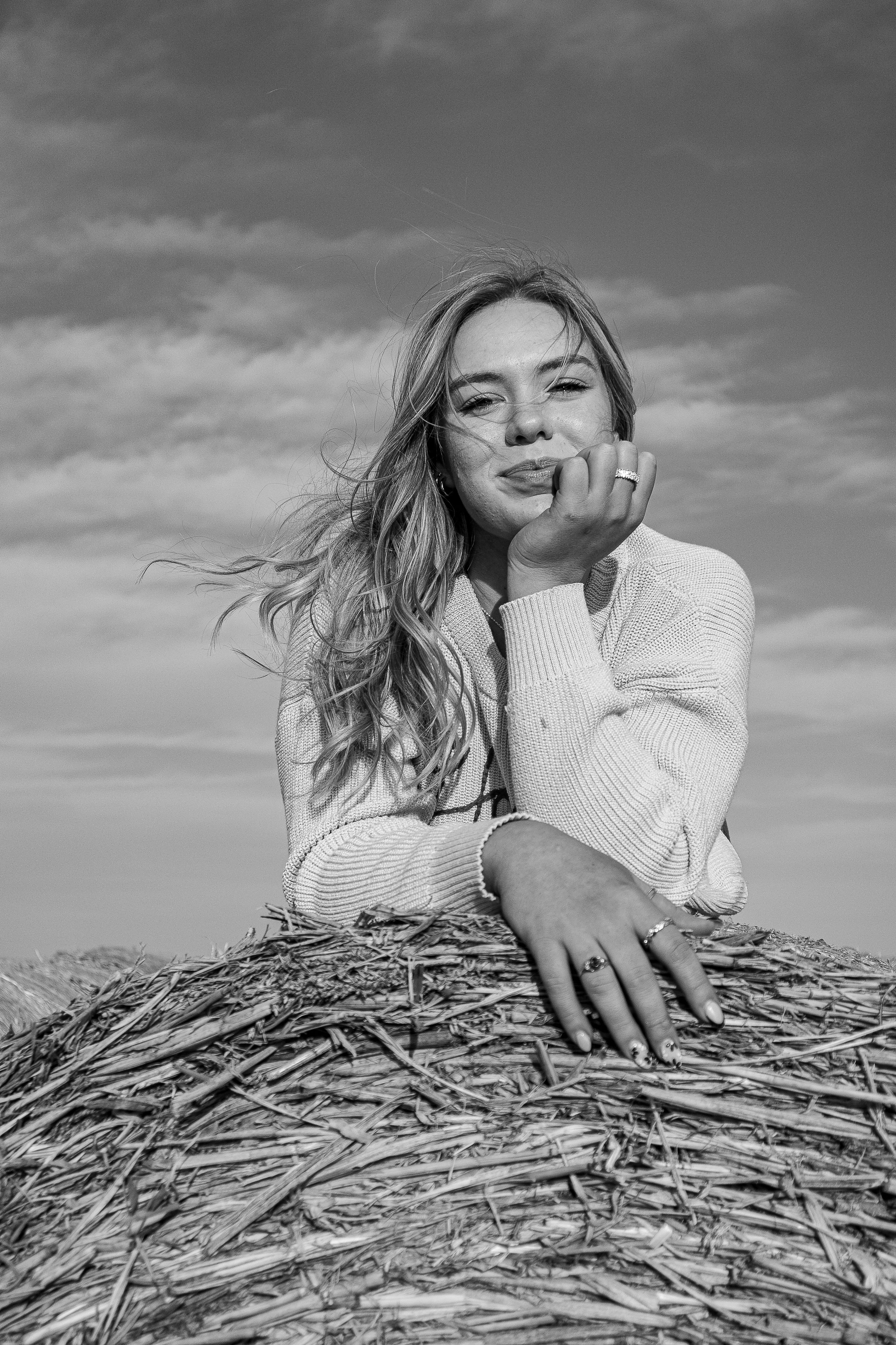 Black and white portrait of person in white sweater posing on hay bale with windswept hair against cloudy sky.
