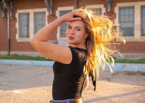 Long blonde hair flows in sunlight as figure in black top poses dramatically against brick building background.