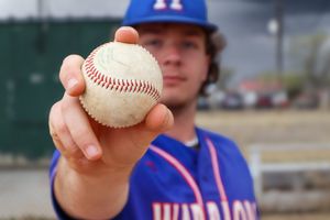 Baseball player in blue uniform holding up a baseball towards the camera.