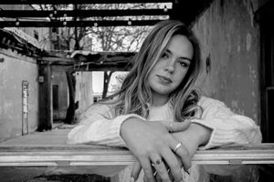 Black and white portrait of someone with long hair leaning on a railing in an industrial setting.
