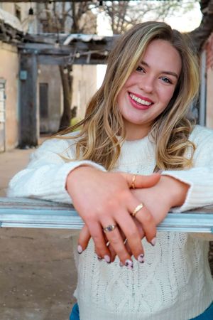 A person in a white cable knit sweater smiling warmly while showing off rings on their hands against an outdoor background.