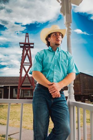 Person in turquoise shirt and cowboy hat standing on porch with red tower in background.