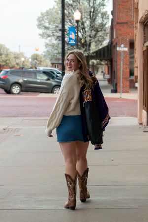 Student wearing cream sweater, skirt and cowboy boots with letterman jacket on downtown sidewalk by brick buildings.