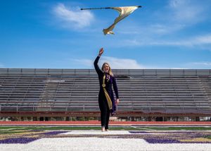 Graduate in black gown tossing their cap in celebration at an empty stadium with blue sky.