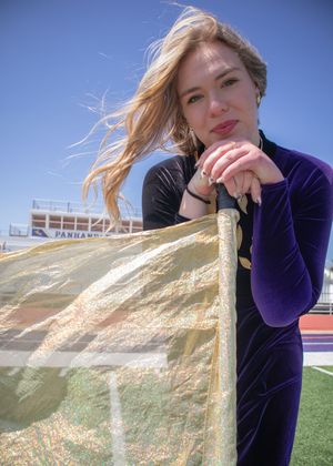 Athletic figure in a navy athletic outfit leans on a pole against a bright blue sky and sports stadium background.