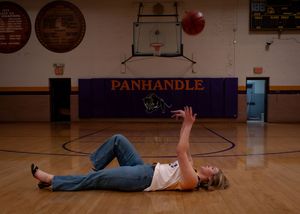 Person lying on basketball court floor practicing throwing red ball at Panhandle gymnasium.