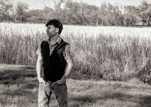 Black and white portrait of figure standing in tall grass field wearing vest and jeans.