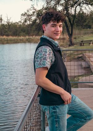 Person in patterned shirt and vest poses by lake railing during sunset.