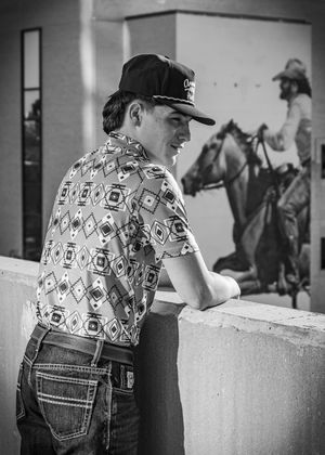 Black and white photo of person in patterned shirt watching horse riders in background.