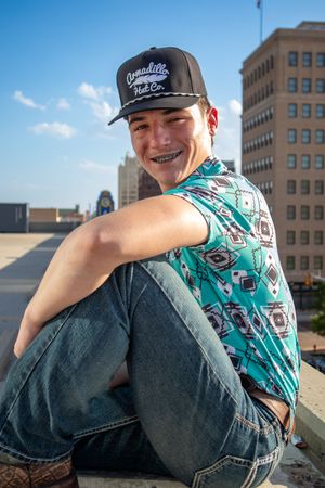 Person in patterned shirt and baseball cap sitting on urban rooftop edge.