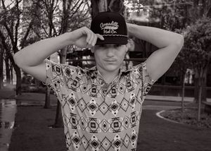 Black and white photo of person adjusting baseball cap wearing geometric patterned shirt.