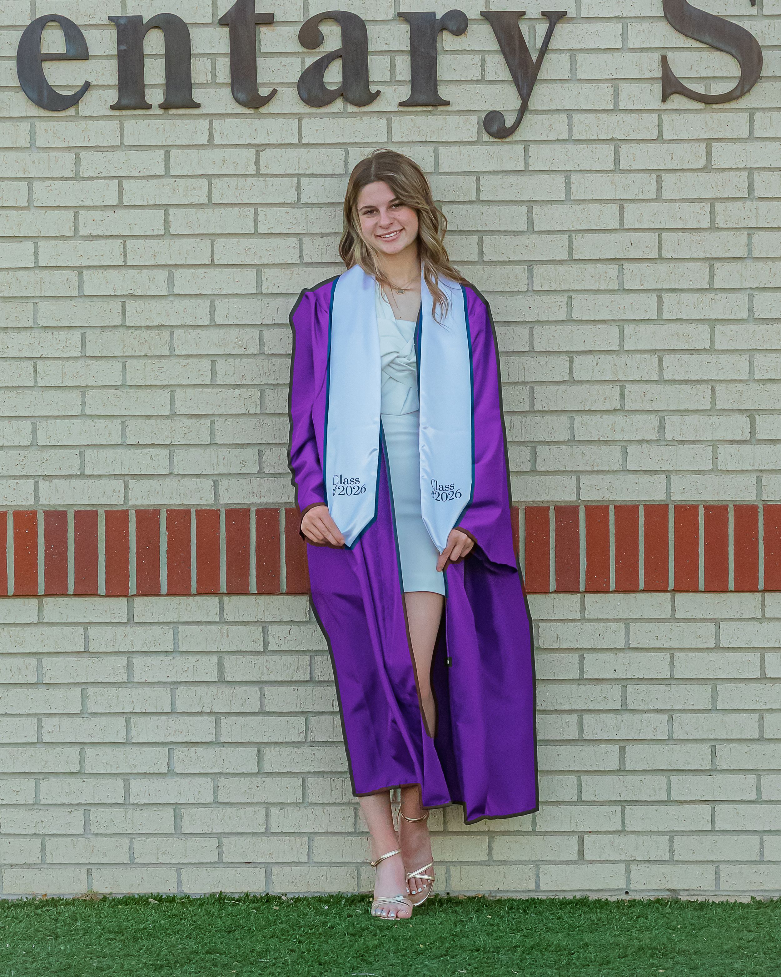 A smiling young woman in a purple graduation gown and white stole poses proudly in front of a brick school wall.