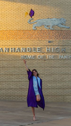 A graduate in purple cap and gown celebrates outside Panhandle High School, Home of the Panthers, raising her diploma joyfully.