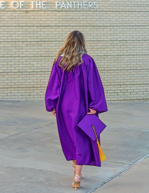 Graduate in purple cap and gown walks toward brick wall engraved with 'Home of the Panthers'.