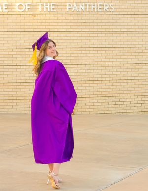 Graduate in purple cap and gown poses in front of brick wall reading Home of the Panthers.