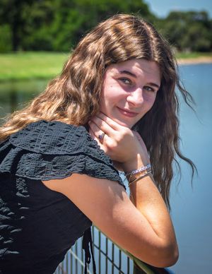 Outdoor portrait of a smiling person with long wavy brown hair wearing a black top leaning against a railing near a lake.