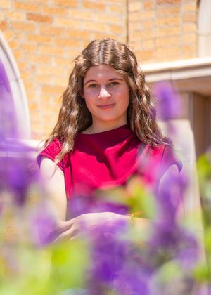 Portrait photo of someone in a bright red top framed by purple flowers in the foreground with a brick wall background.