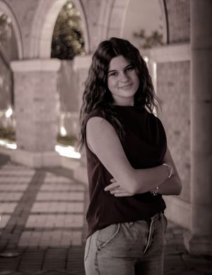 A person wearing a black sleeveless top and jeans poses with arms crossed in an outdoor archway setting.