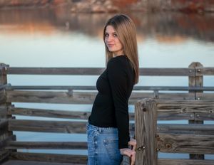 Young woman in black top and jeans poses on wooden dock by calm water at sunset.