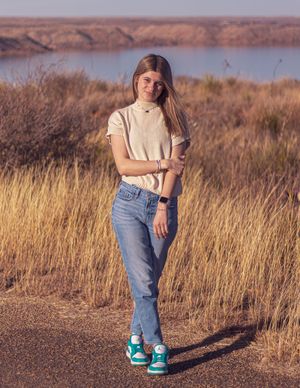 Young woman in jeans and teal sneakers poses on a dirt path with golden grass and a lake in the background.