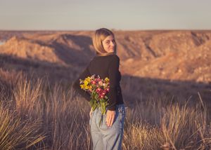 Young woman holding colorful wildflowers behind her back, standing in a scenic desert landscape at golden hour.