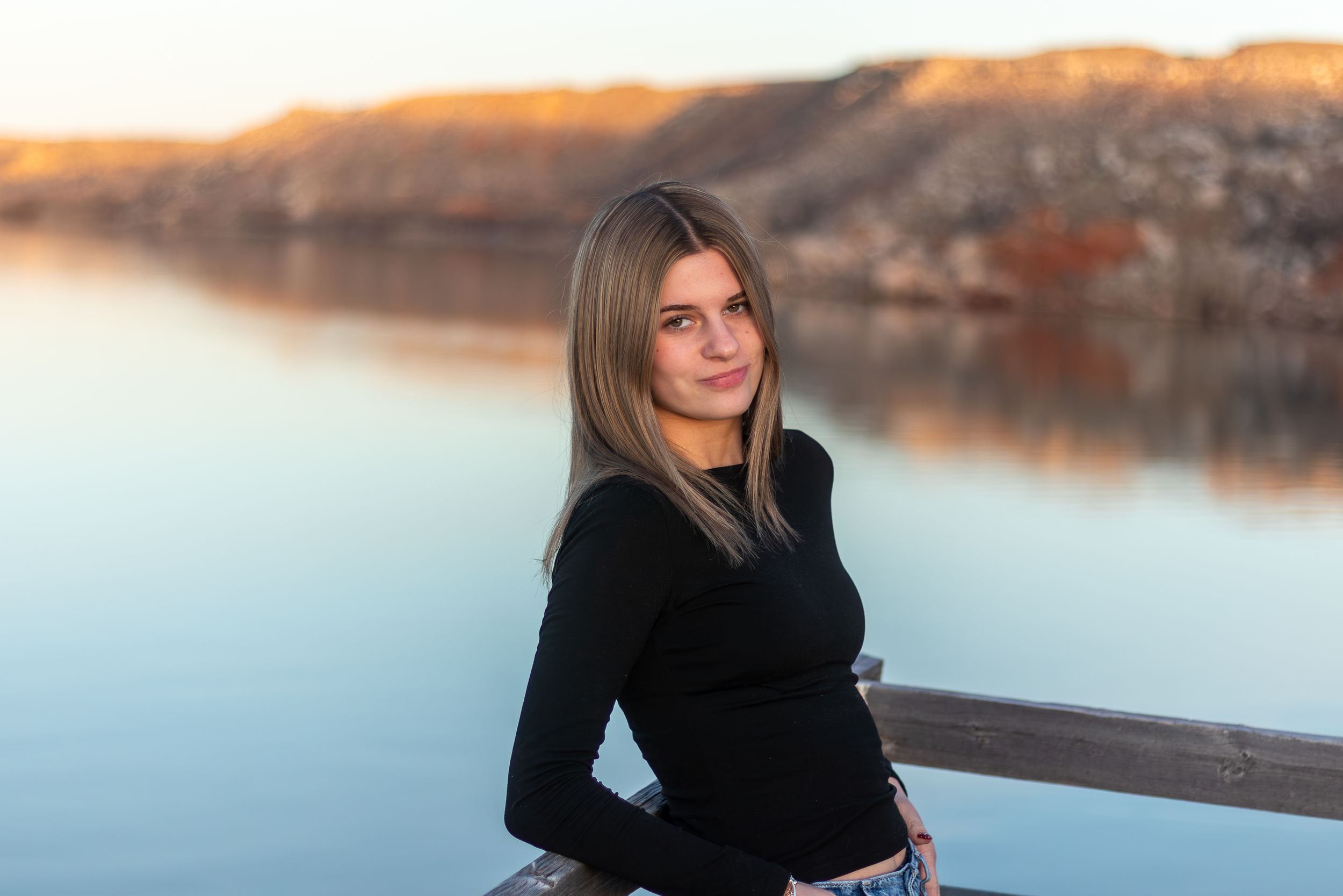 Young woman in black top posing by a serene lake at golden hour with rocky hills in the background.