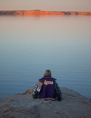 Person in purple varsity jacket sitting on rock overlooking calm lake at sunset with red cliffs in background.