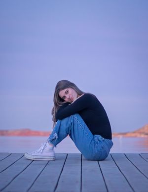 Young woman in black sweater and jeans sits hugging knees on wooden dock at dusk with blue sky backdrop.