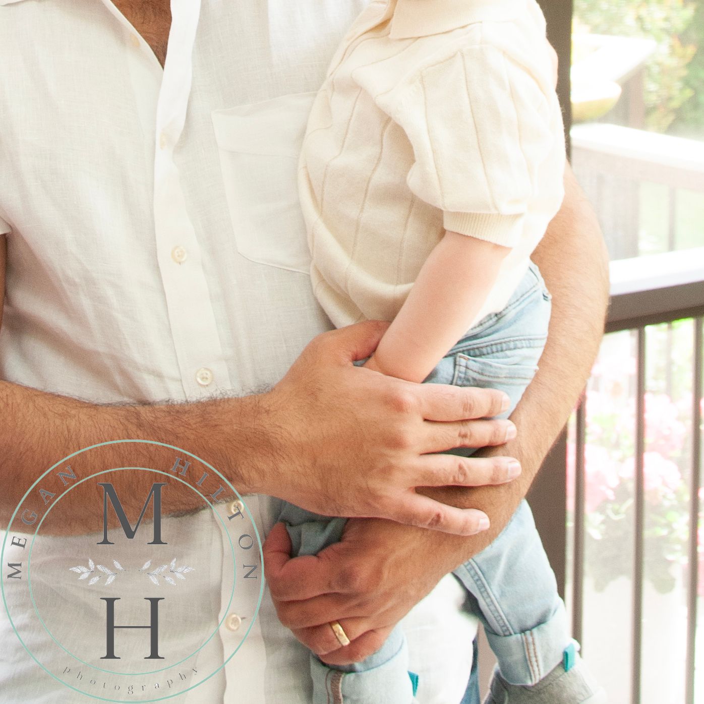 Close-up of hands holding a toddler wearing blue denim, with soft natural lighting through a railing outdoors.