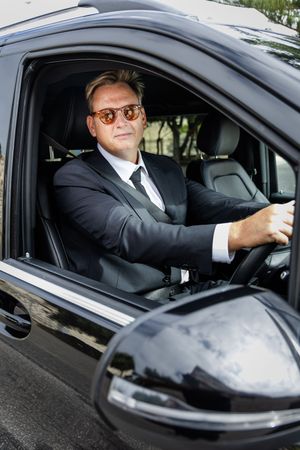 Professional chauffeur in a black suit and sunglasses sits at the wheel of a luxury vehicle.