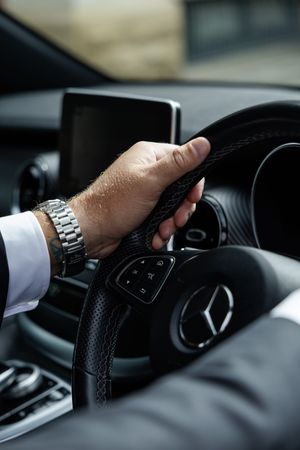 Close-up shot of a person's hand gripping a Mercedes-Benz steering wheel while wearing a silver watch and white sleeve.