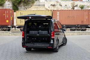 A sleek black Mercedes camper van with a roof rack parked near cargo containers and a concrete wall.