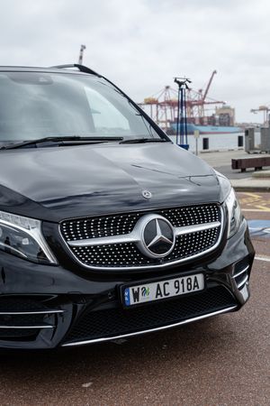 Black Mercedes-Benz V-class van with diamond grille parked at harbor with cargo cranes in background.