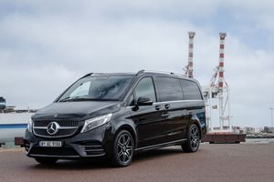 Black Mercedes-Benz V-Class van parked near industrial smokestacks against cloudy sky.