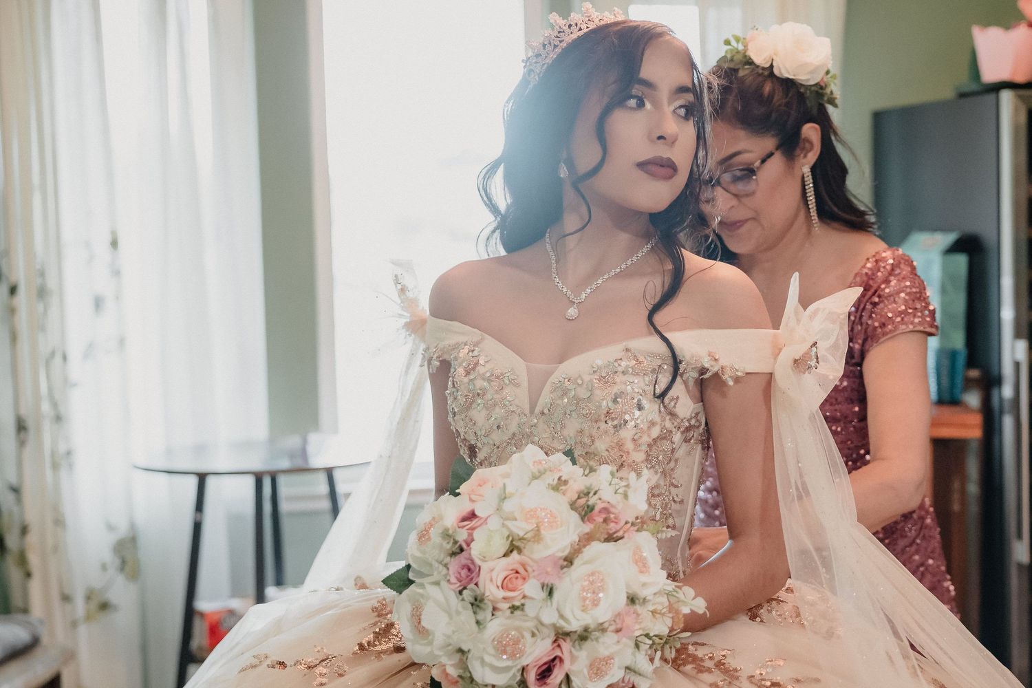 Quinceanera and mother in elegant gowns sit together holding a blush-toned floral bouquet in a sunlit room.