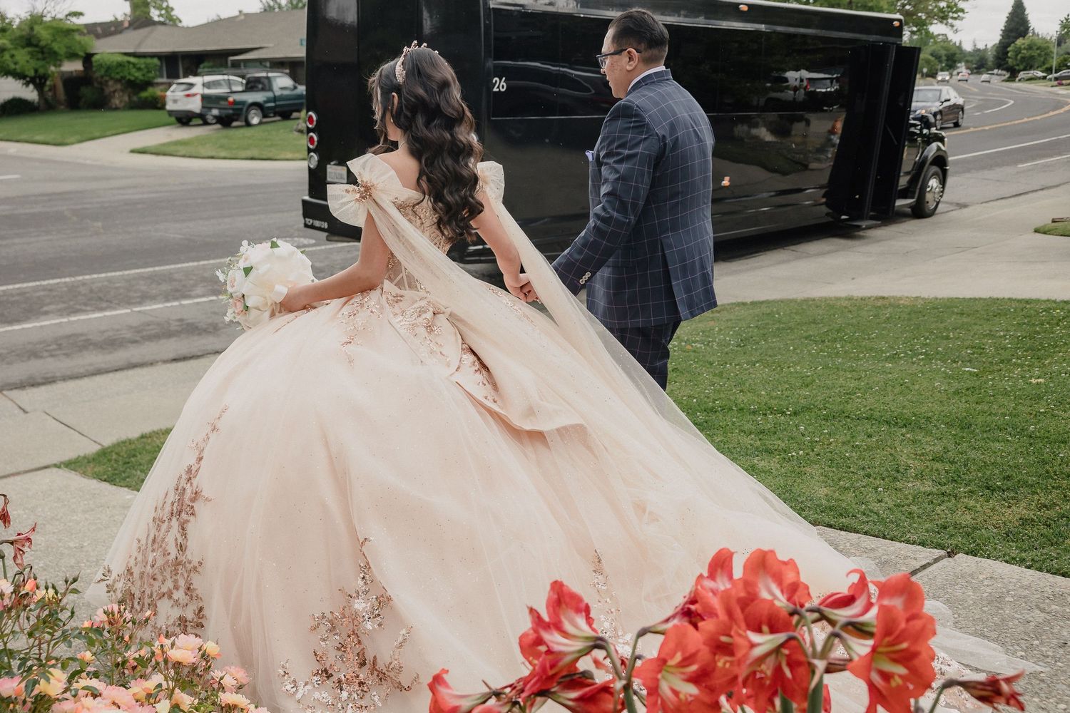 A quinceanera in a blush ballgown and groom enter a black limousine surrounded by red flowers.