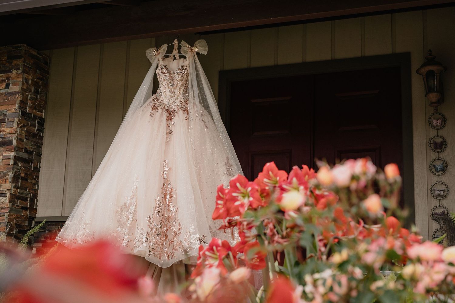 An elegant white quinceanera dress with lace details hangs on display outdoors with red flowers blooming in the foreground.