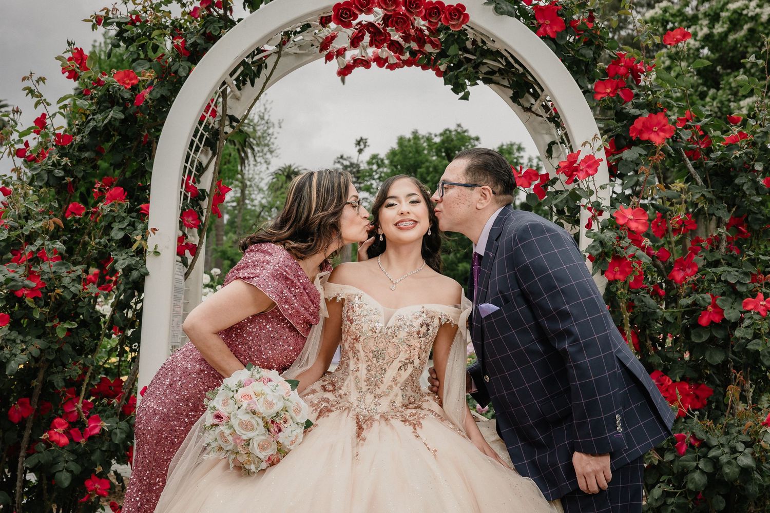 Quinceanera celebration with celebrant under white arch adorned with climbing red roses.