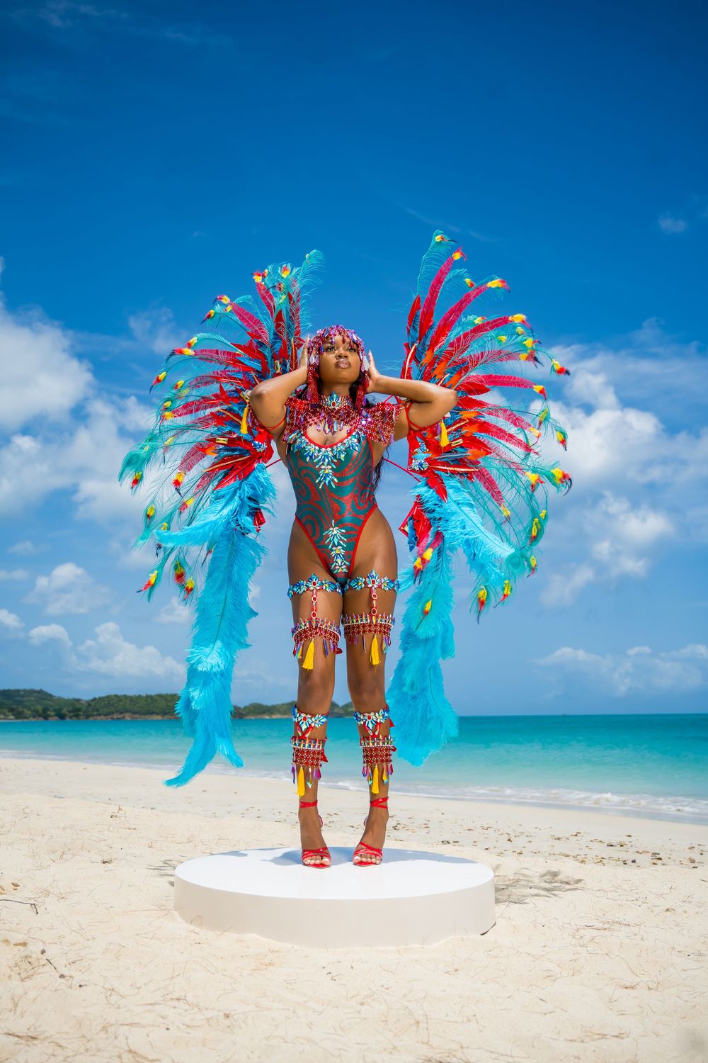 Carnival dancer in turquoise and red feathered costume poses against ocean backdrop.