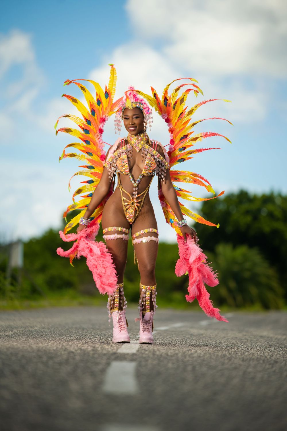 Carnival performer in pink and yellow feathered costume with beaded details poses on road.