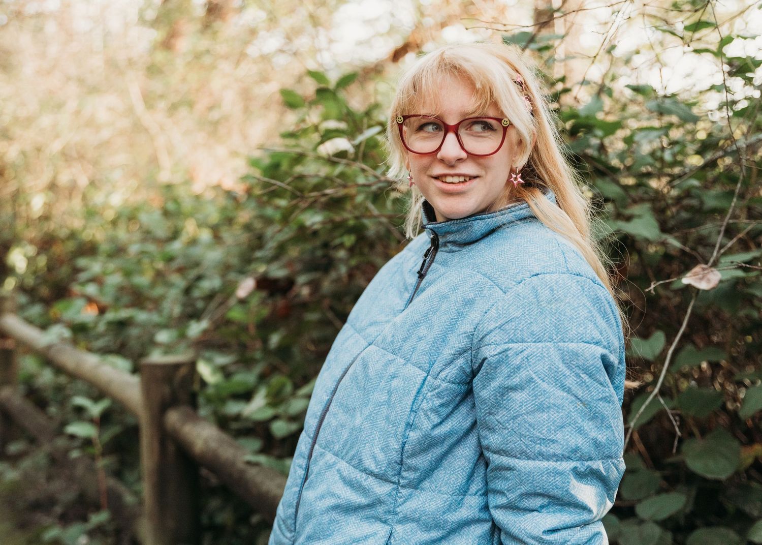 Person wearing glasses and a light blue quilted jacket smiles while standing outdoors near greenery in soft lighting.