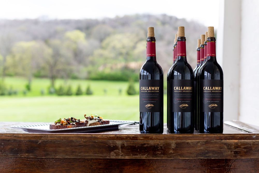 Three bottles of red wine displayed on a wooden surface with a scenic country view in background for a fundraiser event.