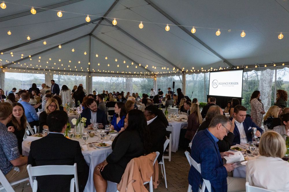 Large group dining under a white tent with string lights at an outdoor fundraiser event.