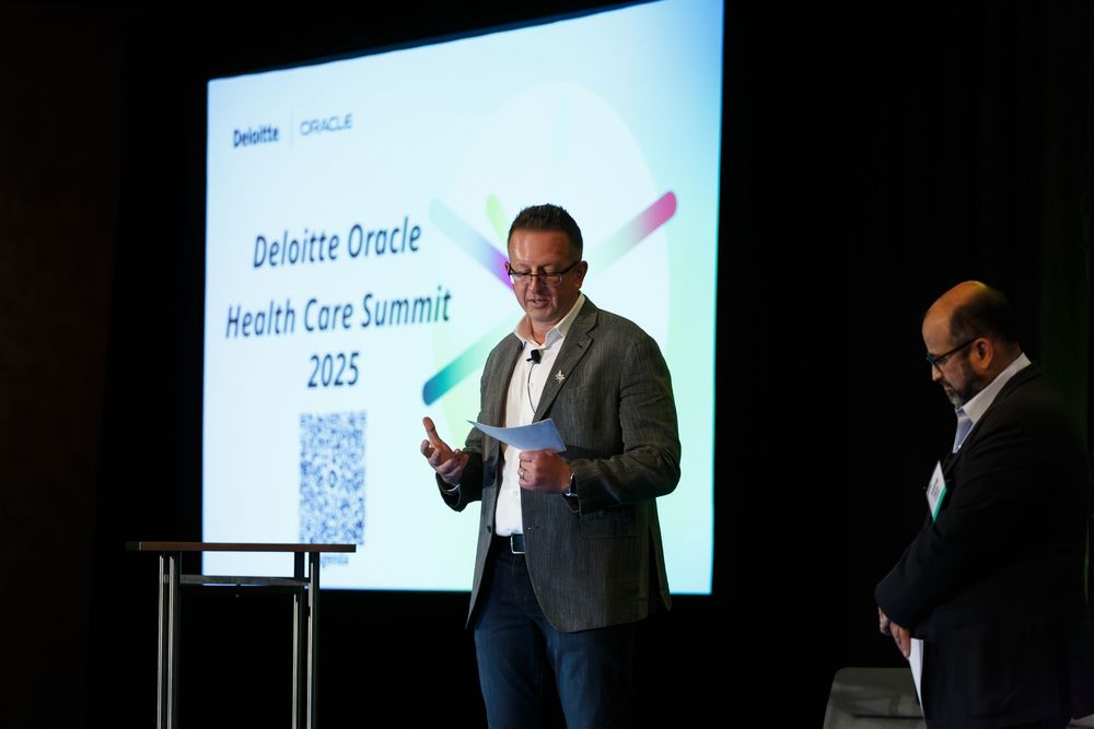 Presenter speaking on stage at a business conference with digital display in background.