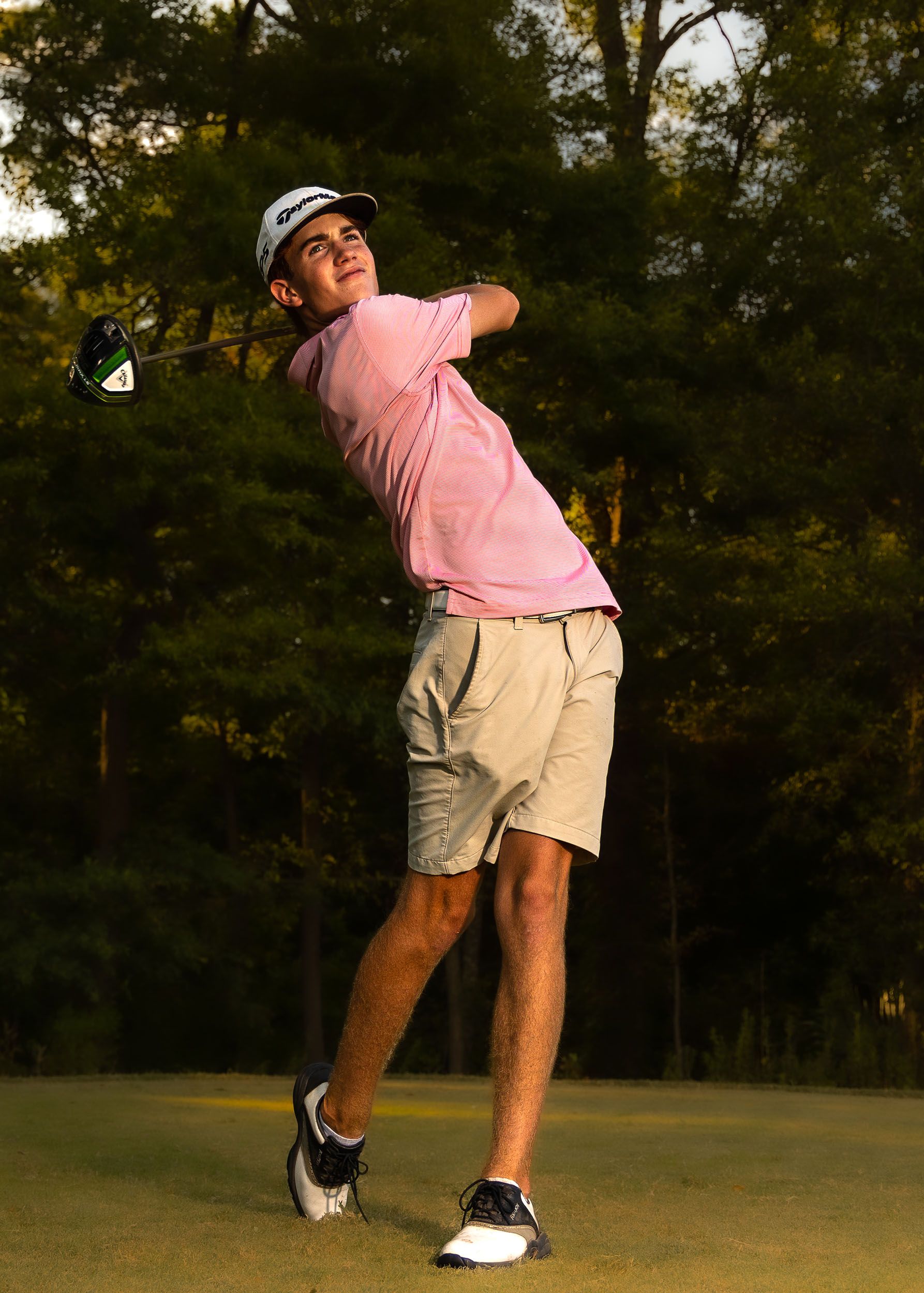 A golfer in a pink polo shirt and khaki shorts takes a swing during golden hour on a golf course.