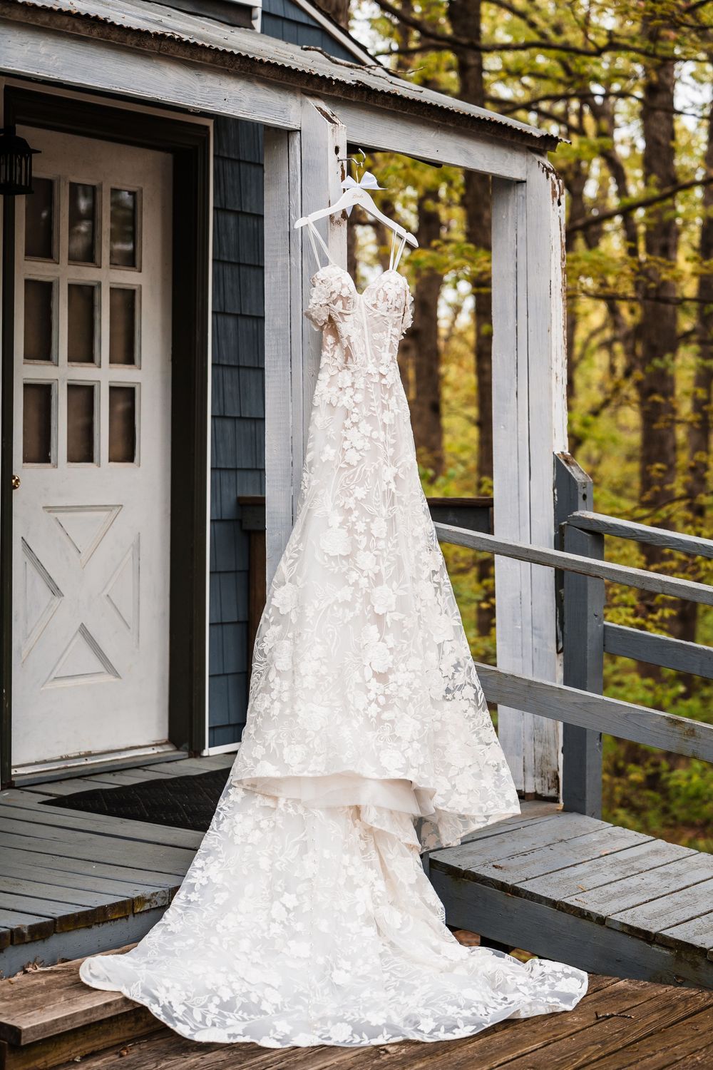 A delicate white lace wedding dress with a long train hangs on display on a rustic cabin porch surrounded by autumn trees.